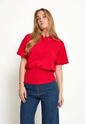 Vrouw met lang blond haar, gekleed in een rood poloshirt met korte mouwen en een blauwe spijkerbroek, staand tegen een witte achtergrond.
