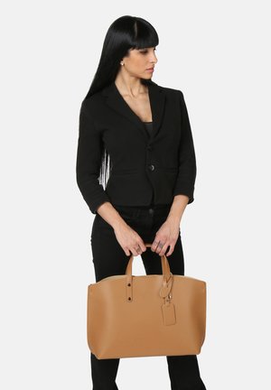 Borsa tote in pelle color tan con due manici superiori, texture liscia e forma rettangolare. Presenta un'etichetta per bagagli e un design minimalista.