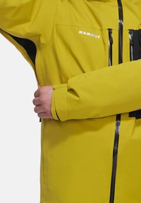 Gelbe wasserdichte Jacke mit schwarzen Akzenten, ausgestattet mit Reißverschlüssen, hohem Kragen und verstellbaren Manschetten, aus strapazierfähigem Stoff gefertigt.