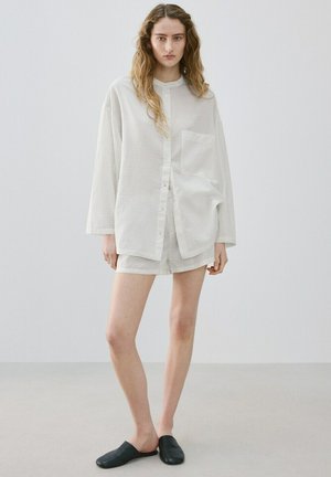 Donna con capelli lunghi e ondulati che indossa una camicia oversize a righe grigio chiaro con bottoni e pantaloncini abbinati, in piedi scalza con scarpe slip-on nere.