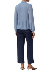 Hellblaue Bluse mit plissiertem Rücken, langen Ärmeln und Kragen, kombiniert mit dunkelblauen Jeans mit weitem Bein und braunen Sandalen mit Absatz.