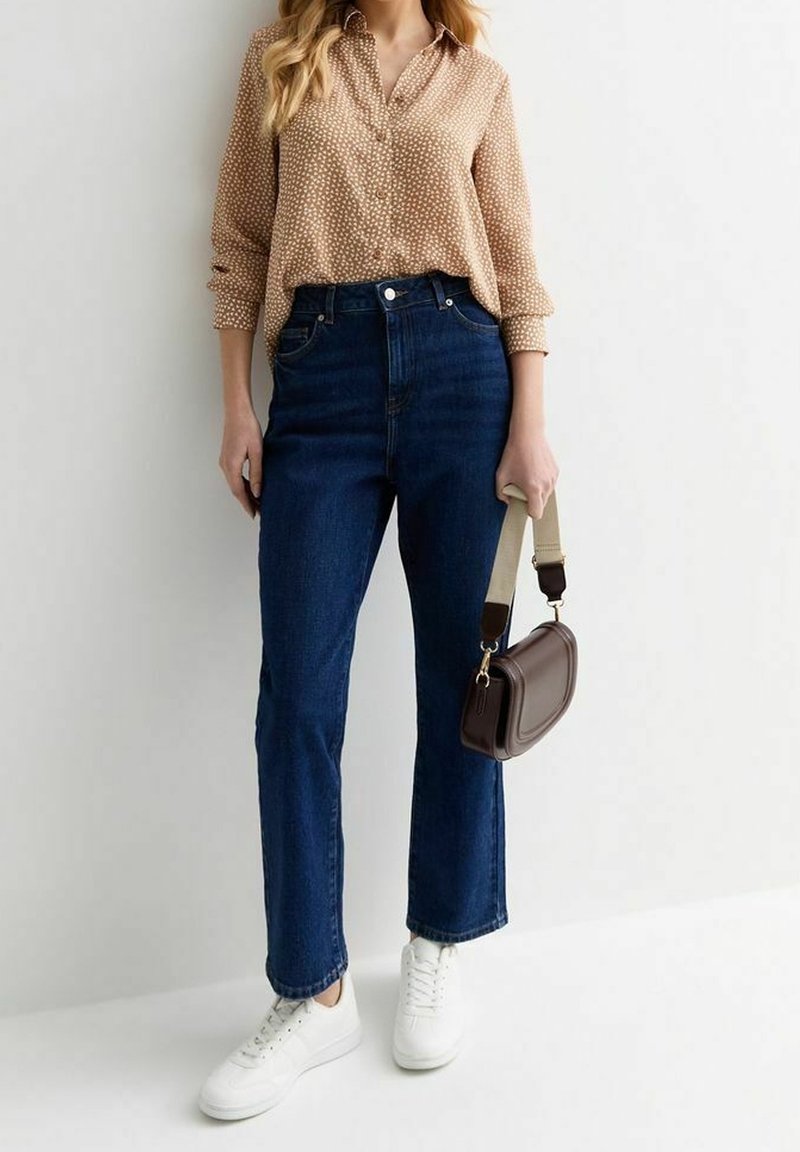 Femme portant un chemisier beige à motifs rentré dans un jean taille haute bleu foncé, baskets blanches, tenant un petit sac à main marron par la lanière.