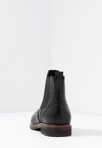 Stivale Chelsea in pelle nera con lati elasticizzati, motivo decorativo a brogue e suola in gomma testurizzata. Finitura lucida con dettagli di cucitura marrone.