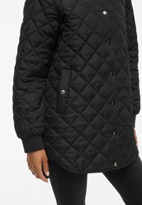 Chaqueta negra acolchada con un patrón de diamantes, botones a presión y bolsillos laterales. Los puños tienen una textura acanalada. Material suave y resistente al agua.