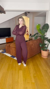 Bordó overall átlátszó hosszú ujjakkal, laza fazonban és V-nyakkal. A modell egy fából készült padlón áll egy textúrázott barna kanapé és egy növény közelében.