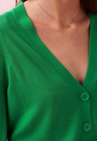 Cardigan verde con scollatura a V, caratterizzato da tre bottoni e una texture liscia. Il materiale a maglia è leggero e morbido.
