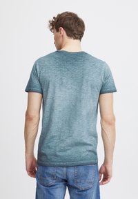 Camiseta corta de manga corta en degradado verde hecha de tela suave. Presenta un cuello redondo clásico y un aspecto ligeramente desvaído. Se lleva con jeans azules.