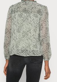 Blouse vert clair en tissu transparent avec un motif tacheté noir. Comprend un col froncé et des poignets élastiqués, avec une coupe décontractée.