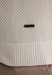 Cremfarbener Strickpullover mit strukturiertem Muster, geripptem Saum und einem zentral platzierten schwarzen Metall-Logoetikett am unteren Rand.