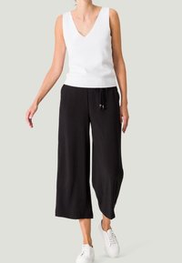 Witte mouwloze V-hals top in combinatie met zwarte wijde broek met een drawstring taille, gedragen met witte sneakers. Neutrale achtergrond.