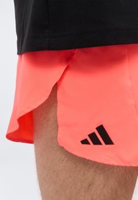 Gros plan d'une personne portant un short de sport corail vif avec un logo Adidas noir et un haut noir partiellement visible.