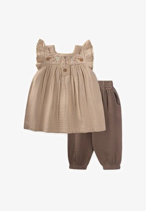 Komplektas apima kreminės spalvos, siuvinėtas palaidines su raukiniais ir sagų apykakle, derinamas su rudiems, siaurėjantiems kelniams, turinčioms elastinius rankogalius.