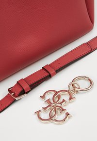 Borsa in pelle rossa con texture e tracolla regolabile. Presenta hardware color oro e un ciondolo con lettere intrecciate.