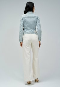 Veste en denim bleu clair avec des détails en boutons et deux poches arrière, associée à un pantalon large crème. Texture lisse, coupe ajustée.