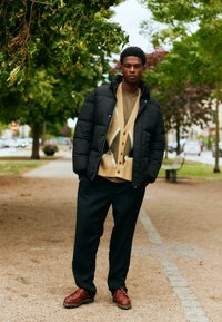 Νεαρός άνδρας που φοράει μαύρο μπουφάν, μπεζ πλεκτή ζακέτα με σχέδια, μαύρο παντελόνι και καφέ παπούτσια, στέκεται σε μονοπάτι σε αστικό χώρο με δέντρα.