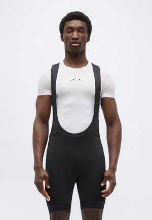 Atleet in een wit gaas Oakley-shirt en zwarte wielerbroek met bretels staand tegen een effen witte achtergrond.