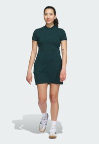 Mörkgrön klänning med kort ärm och subtil textur, slim fit, rundad krage och knälång längd, kombinerad med vita sneakers och strumpor.