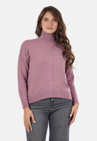 Maglione a collo alto di colore viola chiaro con orlo e polsini a coste, realizzato in un materiale morbido, con un semplice dettaglio di cucitura sul davanti. Indossato con jeans scuri.
