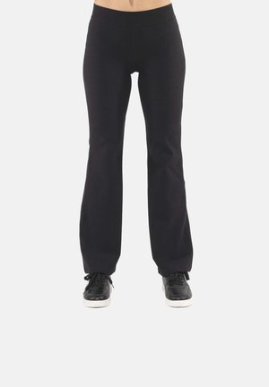 Personne portant un pantalon évasé noir et des baskets noires, debout devant un fond clair uni.