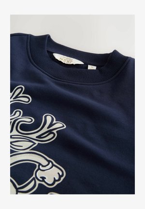 Tummansininen collegepaita, jossa on ribbaus kaula-aukossa. Ominaisuutena valkoinen graafinen design sarvista. Tunniste näkyvissä ja siinä on brändin nimi ja koko.