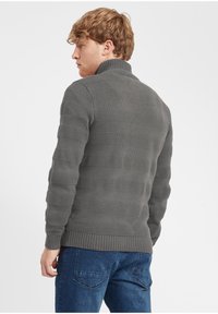 Suéter gris de punto con rayas de textura elevada, cuello alto y puños acanalados. Llevado con jeans azules.