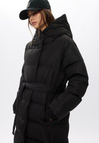 Giacca a secchiello nera con vita cinturata, colletto alto e cappuccio. Presenta una trama trapuntata e tasche laterali, realizzata in un materiale elegante e liscio.