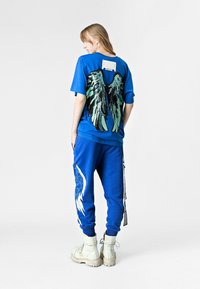 Camiseta azul extragrande con un diseño de alas en verde y negro en la espalda, combinada con joggers azules a juego que presentan detalles de alas blancas y botas.