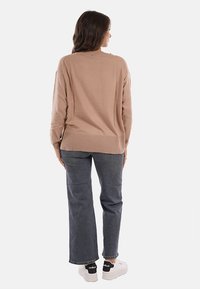 Donna in piedi di spalle, che indossa un maglione lungo color sabbia, jeans dritti grigio scuro e sneaker bianche con dettagli neri sul tallone.