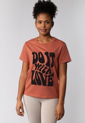 Femme aux cheveux bouclés, de face, portant un T-shirt couleur rouille avec un texte noir indiquant « FAIS-LE avec AMOUR ».