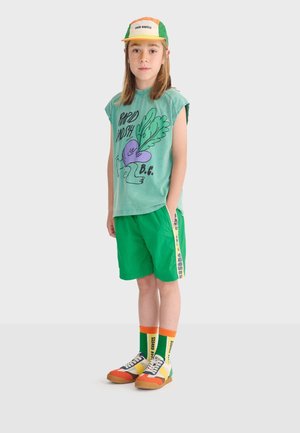 Niño con camiseta sin mangas con diseño gráfico verde, pantalones cortos verdes con franja lateral, zapatillas multicolores, calcetines altos y coloridos, y una gorra amarillo-naranja-verde.