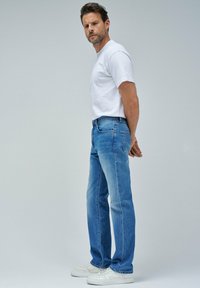 Vaqueros de denim azul con un corte recto, lavado medio y detalle en los bolsillos traseros, combinados con una camiseta blanca ajustada y zapatillas blancas.