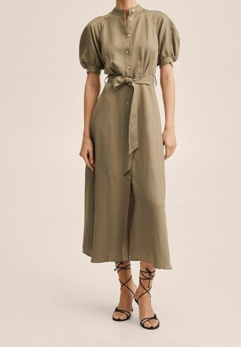 Femme portant une robe midi beige boutonnée avec des manches bouffantes, une ceinture nouée à la taille et des sandales noires à talons hauts et à brides.