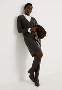 Vestido gris de punto con escote en V y acentos de encaje en los puños, combinado con botas de cuero marrón hasta la rodilla y un bolso marrón oscuro.