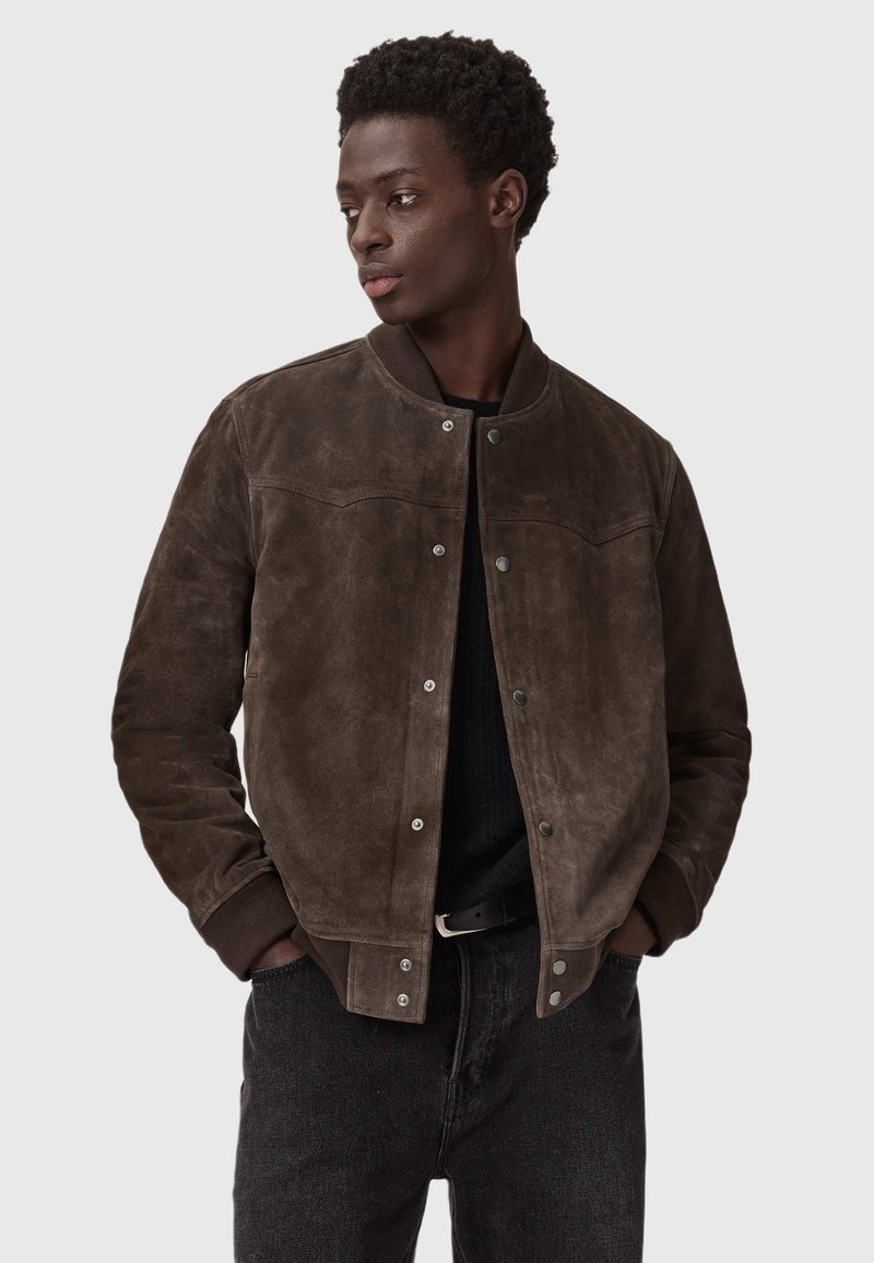 Brązowa skórzana kurtka bomberka z prążkowanym kołnierzem i mankietami, z zapięciem na napy i dopasowanym krojem. Noszona na czarnym swetrze.