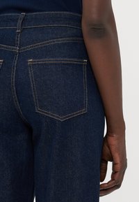 Mørkeblå denimbukser med høy midje, med oransje sømmer og to baklommer. Stoffet har en teksturert og robust utseende.