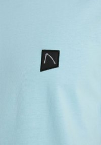 Hellblaues Kurzarm-T-Shirt mit einem kleinen schwarzen Patch und einem weißen geometrischen Design. Glatte Textur mit taillierter Silhouette.