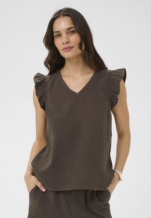 Femme aux longs cheveux foncés portant un haut marron foncé sans manches à décolleté en V avec des épaules volantées et un pantalon assorti, les mains dans les poches.