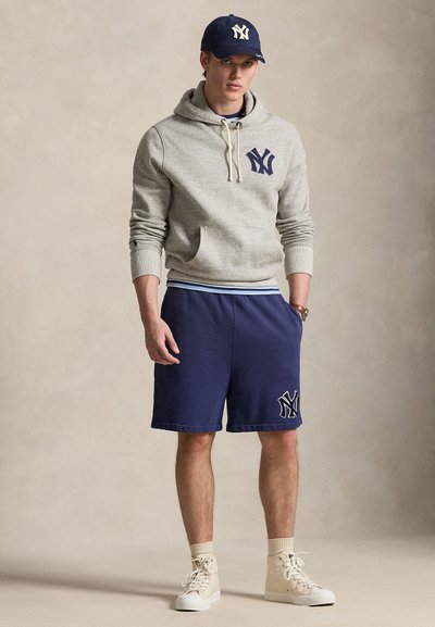 Siva jopa s kapuco in sprednjim žepom ter mornarsko modrim logotipom NY, združen s kratkimi hlačami v isti barvi z enakim logotipom. Svetli supergi zaključujejo videz.