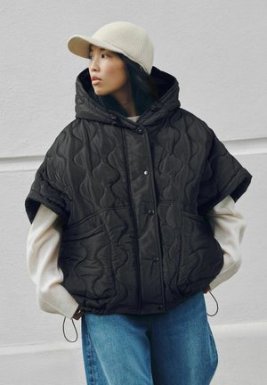 Kvinde, der kigger til siden iført en sort quiltet hætteponcho, hvid langærmet trøje, blå jeans og en beige strikket hue.