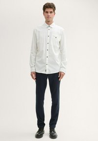 White, long-sleeve button-up shirt with a pocket and black buttons, paired with navy trousers and black shoes, standing against a light background.