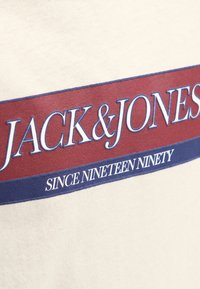 Tessuto color crema con il testo "JACK & JONES" in bianco su un rettangolo rosso e "SINCE NINETEEN NINETY" in bianco su un rettangolo blu sotto.