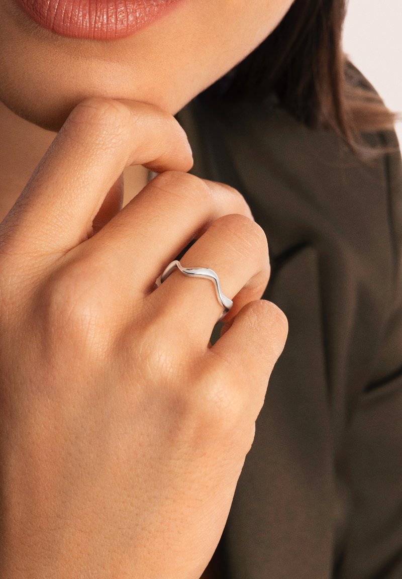 Anillo de ola de plata con un diseño curvado y suave; la banda es delgada y brillante, reposando en una mano con un tono de piel natural.