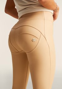 Leggings beige aderenti con tessuto testurizzato, cuciture curve lungo i fianchi e un piccolo logo metallico sulla tasca posteriore.