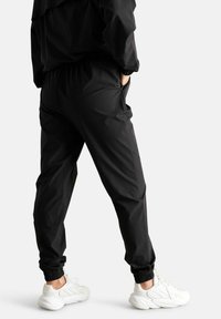 Pantaloni da jogging neri in tessuto elastico con vita e polsini, dotati di tasche laterali e un tessuto leggero e liscio. Indossati con sneakers bianche.
