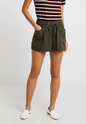 Femme portant un short vert foncé à cordon avec des poches avant, un t-shirt rayé bordeaux et blanc, et des chaussures blanches sans lacets, debout sur un fond uni.