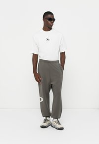 Hvid kortermet t-shirt med logo, grå afslappede bukser med elastisk talje, og beige sneakers. Model med mørke solbriller.