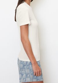 Kvindelig iført en tætsiddende hvid T-shirt med korte ærmer og lyseblå denim shorts med kænguru-logo mønster, står sidelæns mod en hvid væg.