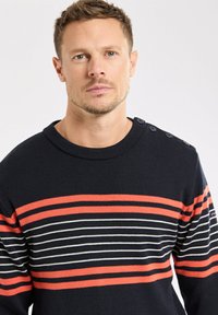 Homme aux cheveux courts portant un pull bleu marine avec des rayures horizontales rouges et blanches et des boutons à l'épaule, se tenant face à un fond blanc.