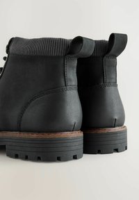 Ghete scurte din piele neagră, cu un guler din material tricotat cu nervuri, prevăzute cu două tăvițe pentru a facilita îmbrăcarea, talpă de cauciuc și cusături maro contrastante de-a lungul bazei.