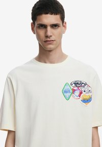 T-shirt in cotone crema con maniche corte, caratterizzata da colori vivaci e toppe ricamate di varie forme e design sul petto.